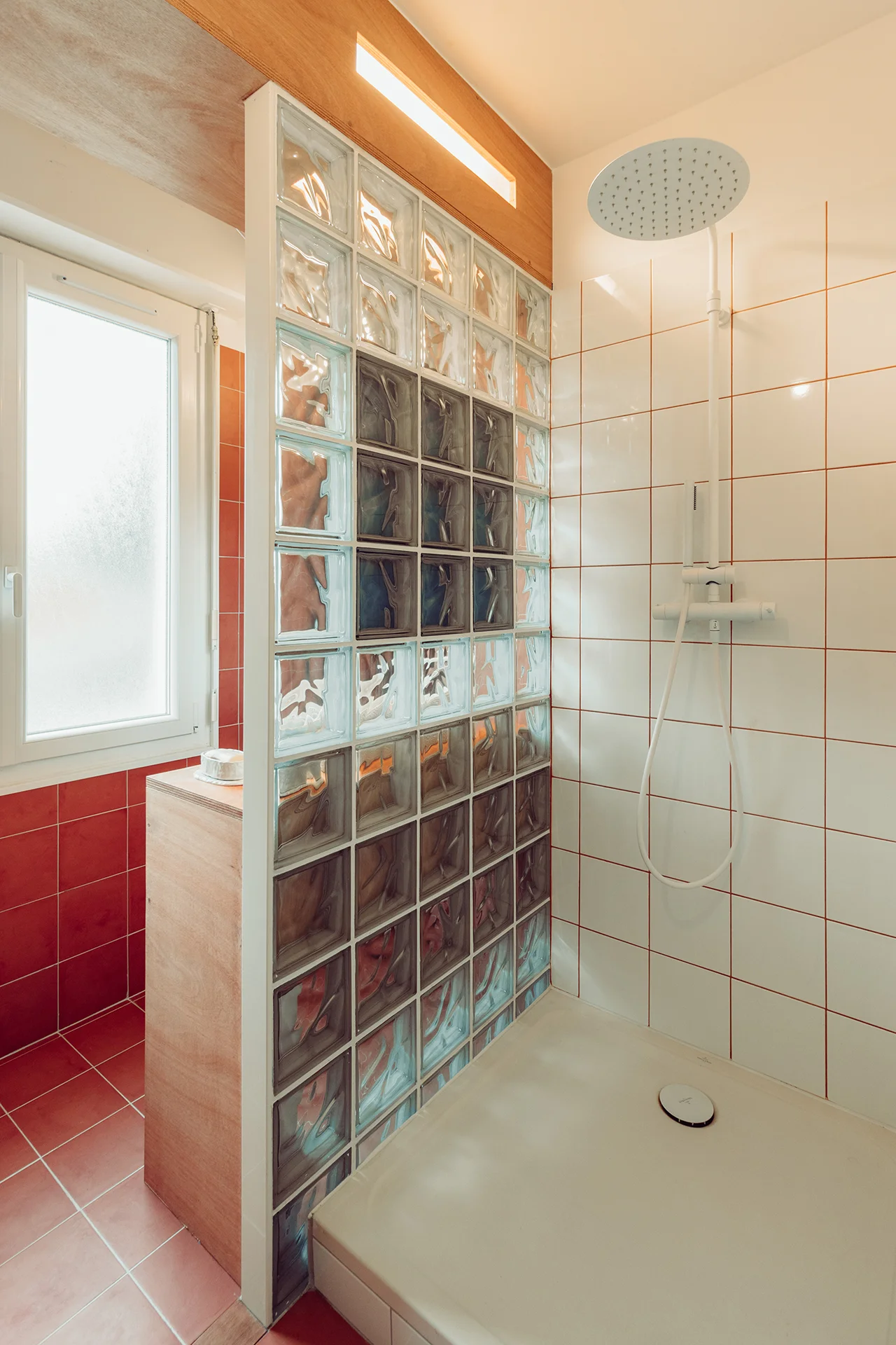 Détail partie douche en négatif du reste (carrelage blanc joints terracotta), colonne de douche blanc mat, éclairage intégré à la douche depuis le caisson en bois